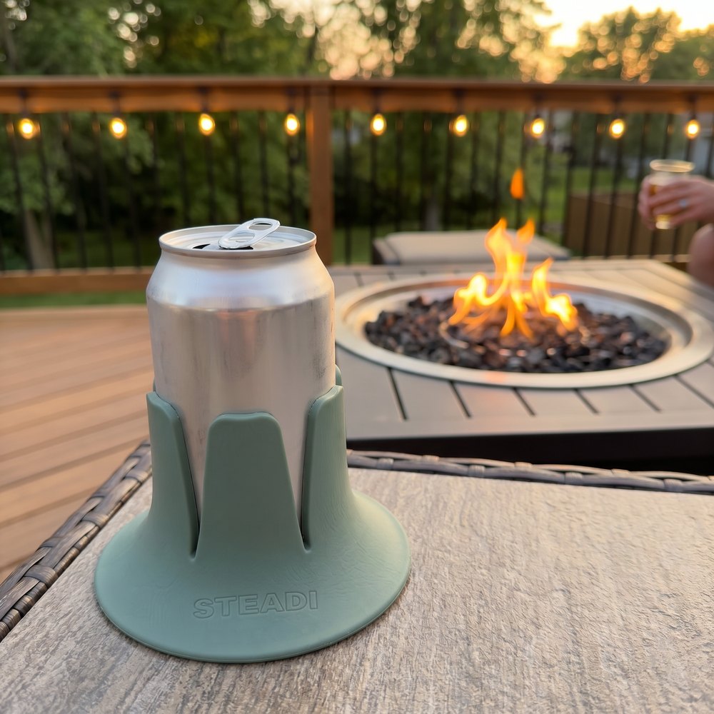 A can standing in a sage Steadi on a deck rail at dusk, a fire and string lights behind.